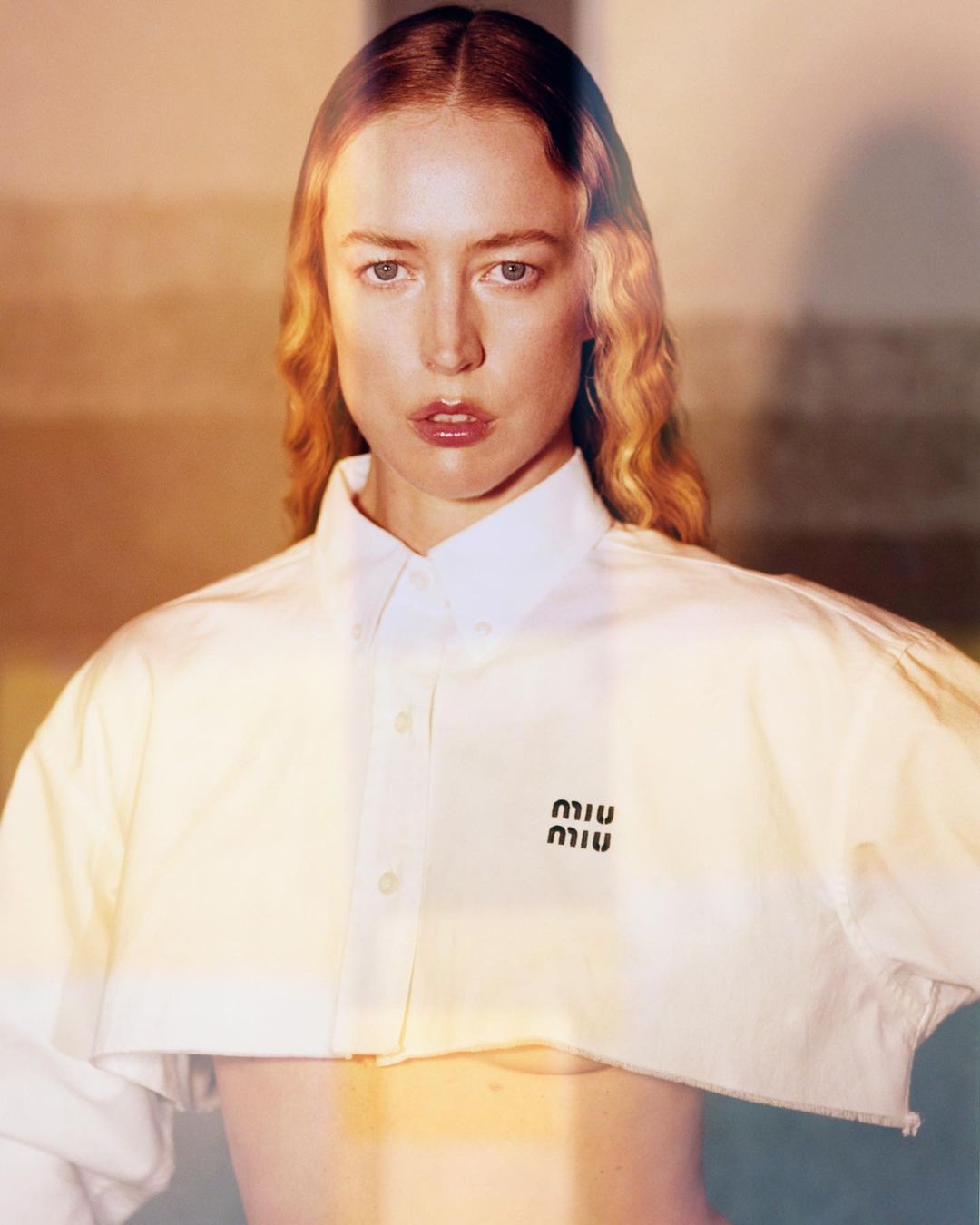 A woman poses wearing a cropped white button-down shirt with a Miu Miu logo, illuminated by a soft, warm light in a studio setting.