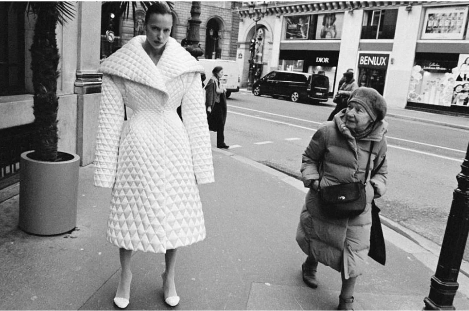 A fashion model poses on a city street wearing a quilted white coat, with an elderly woman walking in the background in black and white.