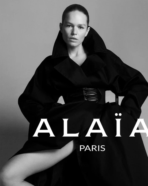 Black and white fashion portrait of a woman wearing a black Alaïa coat with a large collar and a belt, posing in front of a grey background.