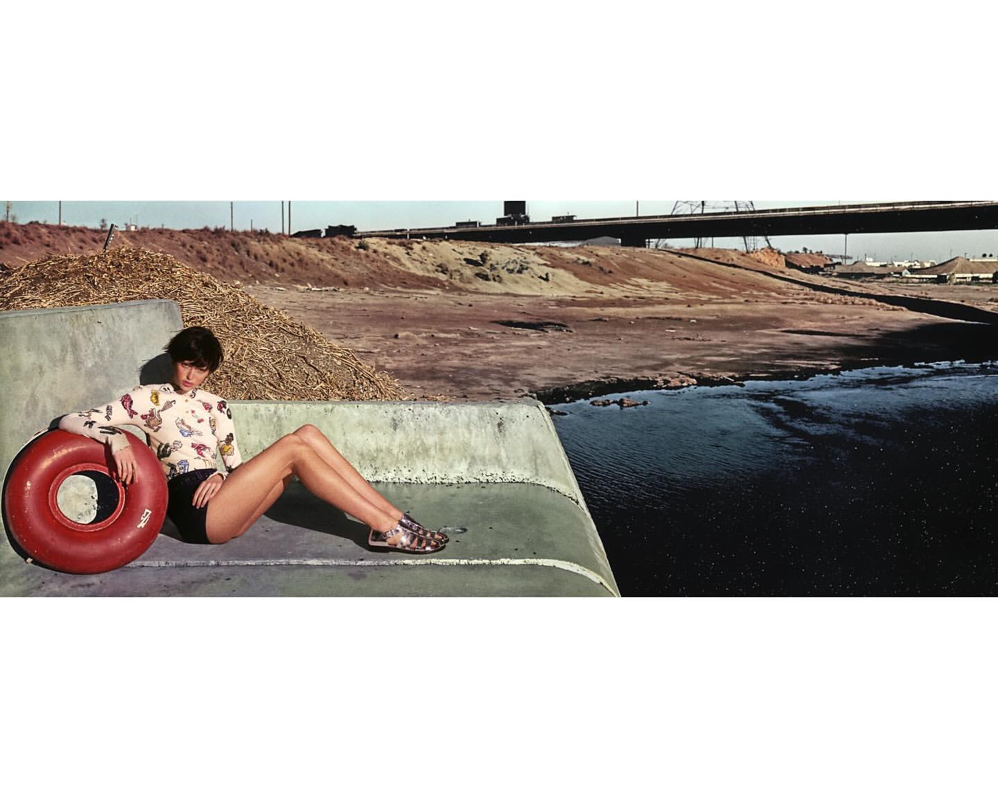 A woman in black shorts and printed top poses with a red inflatable tube on a concrete structure, with a river and highway in the background.