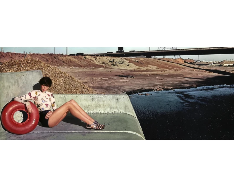 A woman in black shorts and printed top poses with a red inflatable tube on a concrete structure, with a river and highway in the background.