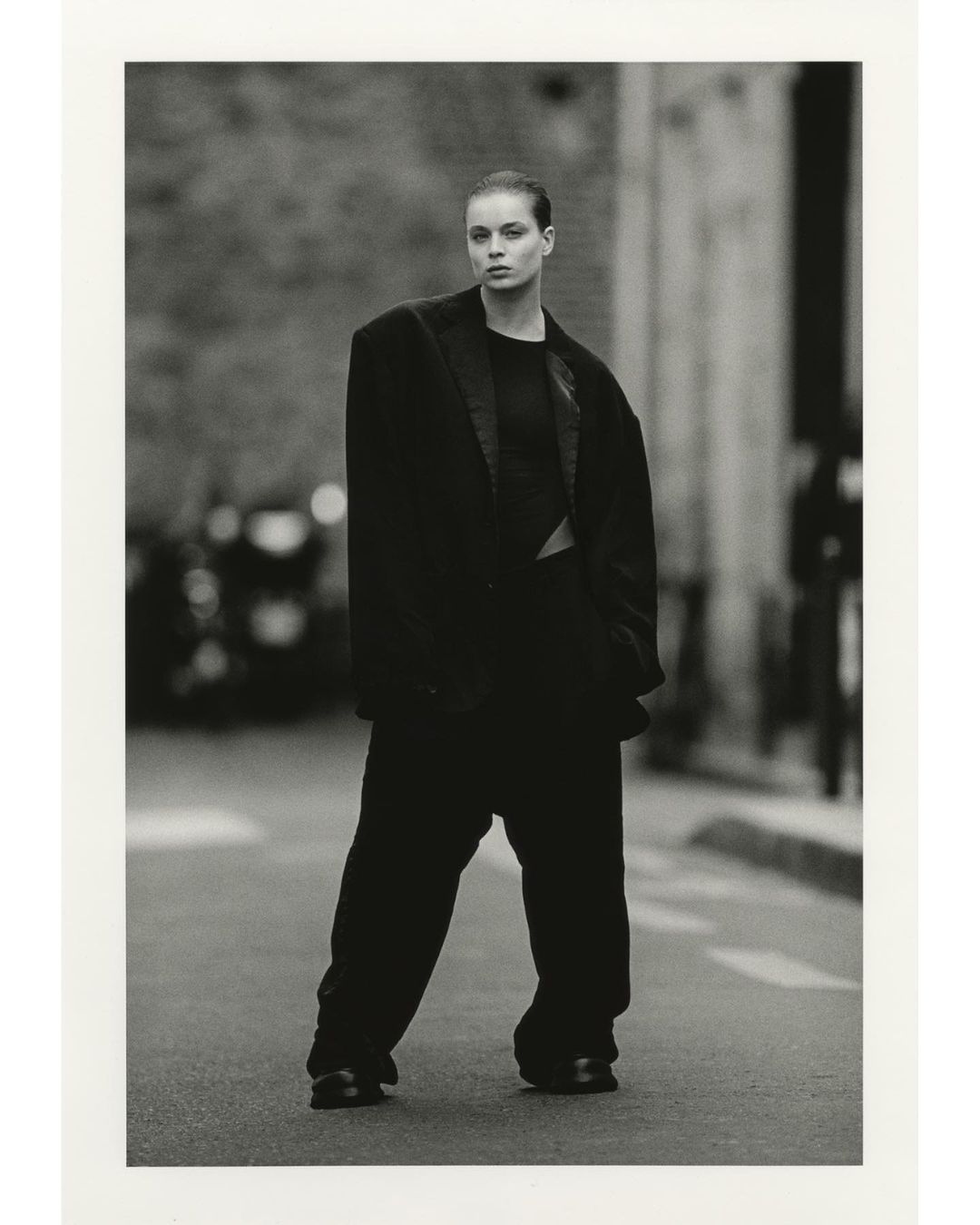 A fashion shot of a woman in an oversized suit standing on a city street, rendered in black and white with a shallow depth of field.