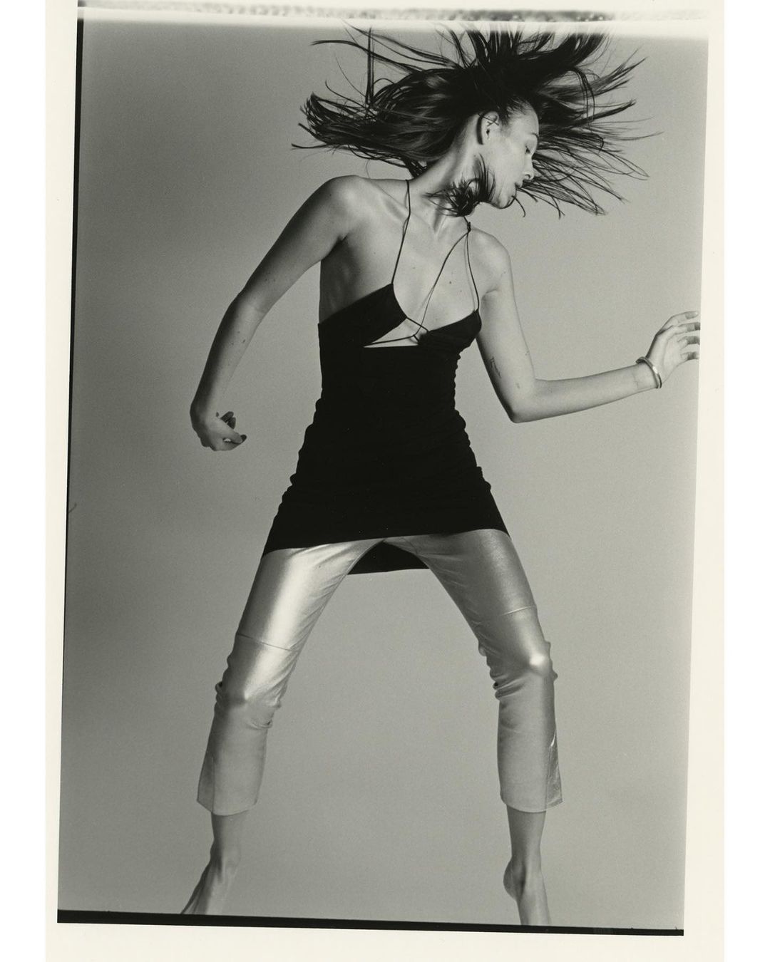 A woman with dark hair and silver pants is in motion against a gray background, wearing a black dress in a fashion-style photograph.