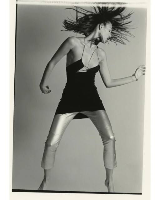 A woman with dark hair and silver pants is in motion against a gray background, wearing a black dress in a fashion-style photograph.