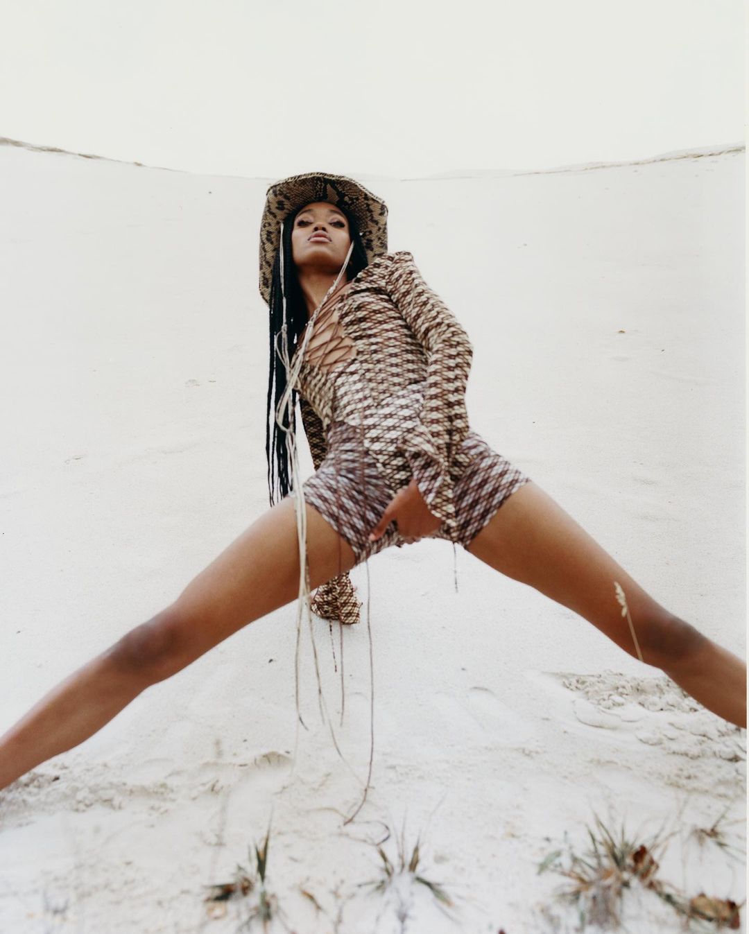 Fashion shot of a woman with braids in a patterned outfit and cowboy hat striking a pose against a neutral dune backdrop.