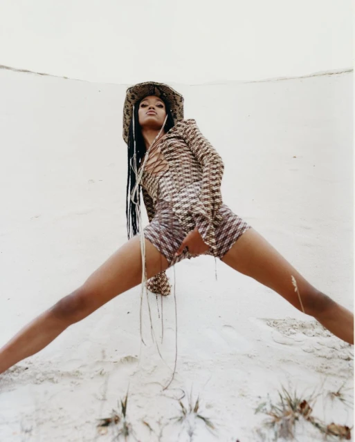 Fashion shot of a woman with braids in a patterned outfit and cowboy hat striking a pose against a neutral dune backdrop.