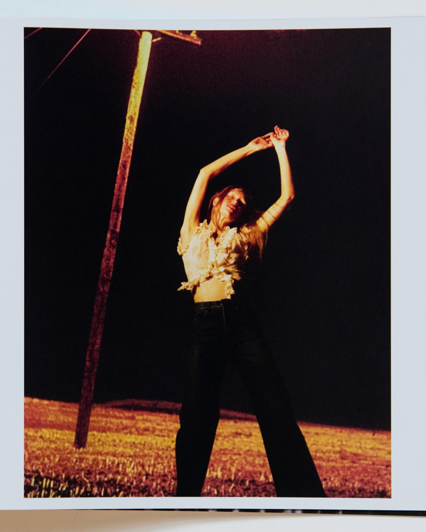 Woman poses with arms raised next to a weathered utility pole at night, illuminated by a warm, orange glow against a black backdrop.