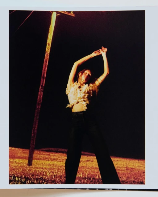 Woman poses with arms raised next to a weathered utility pole at night, illuminated by a warm, orange glow against a black backdrop.