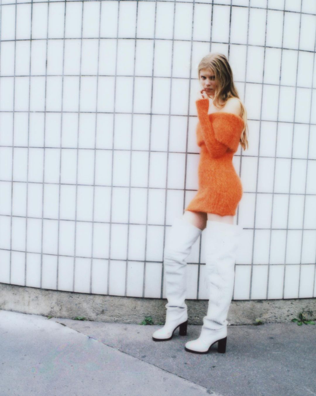 Blonde woman in orange dress and white boots poses against a white tiled wall. Fashion photography with natural light. Shoulder-length blonde hair.