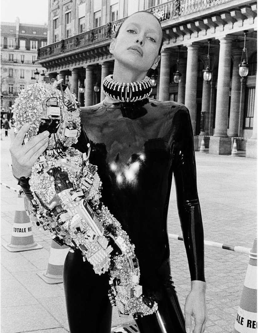 Fashionable woman in shiny black catsuit with a silver embellished arm piece, standing in an urban environment with classic architecture.
