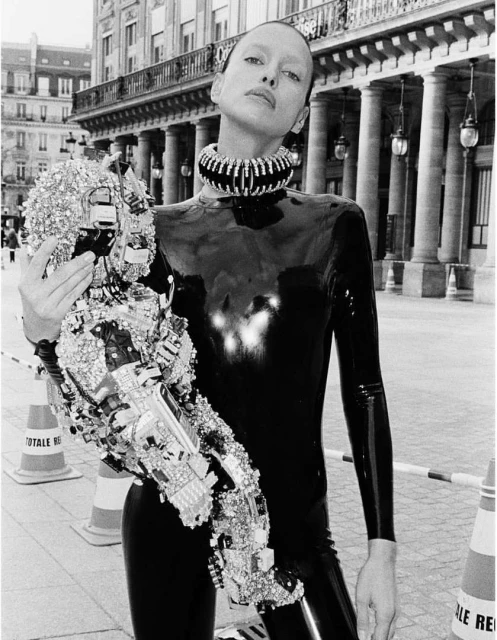 Fashionable woman in shiny black catsuit with a silver embellished arm piece, standing in an urban environment with classic architecture.