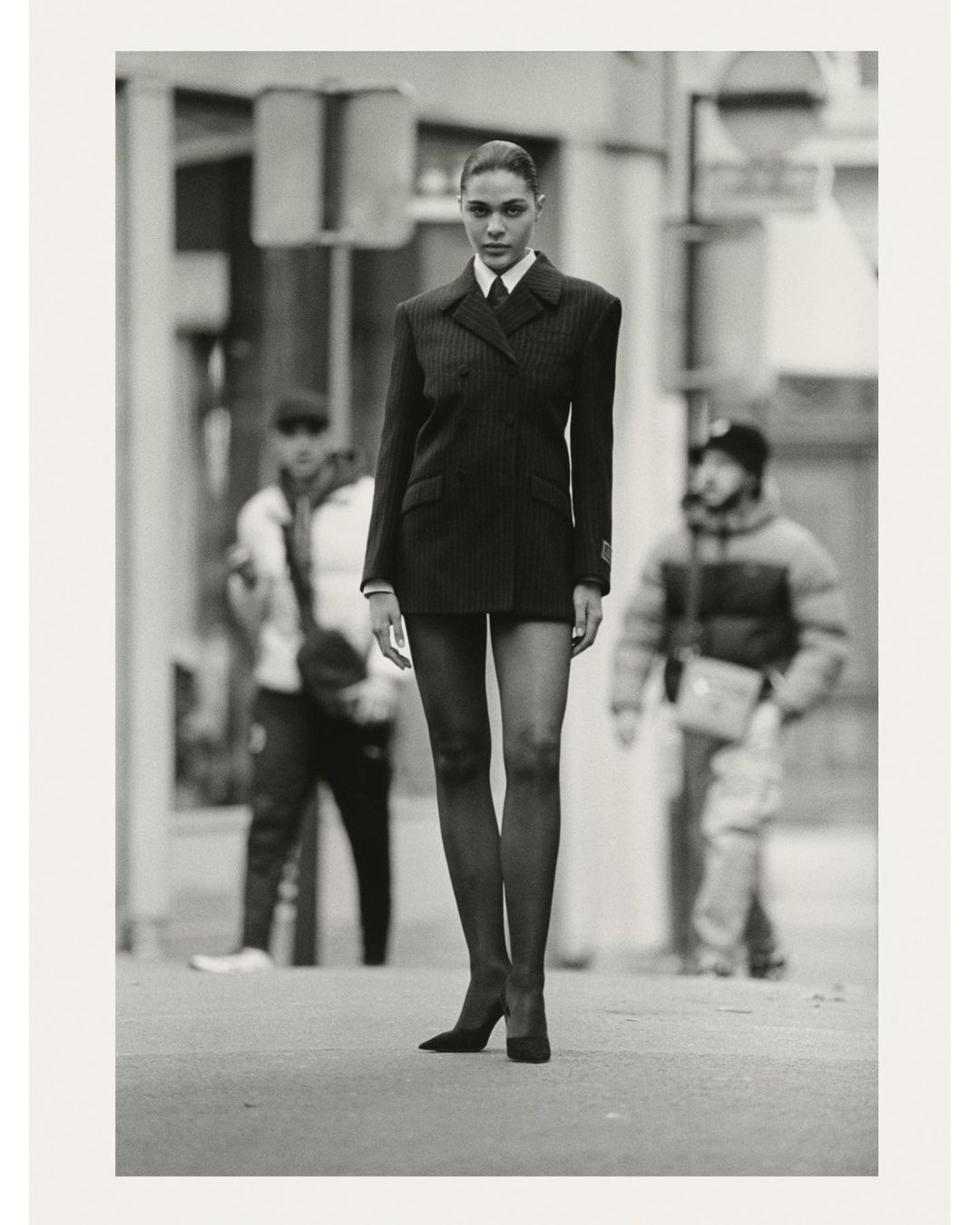 Fashionable woman in suit walks on city street. She has long legs and wears heels. Two blurred people in the background.