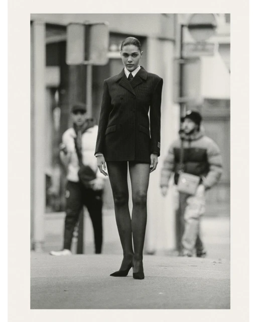 Fashionable woman in suit walks on city street. She has long legs and wears heels. Two blurred people in the background.