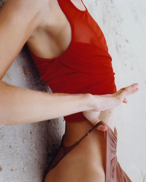 Close-up studio shot of a slender adult woman wearing a red tank top and brown bottoms, standing near a textured white surface.