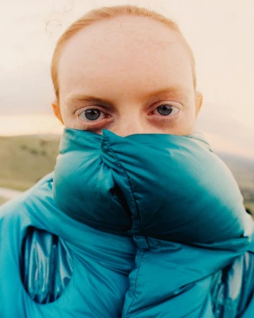 Close-up portrait of a person with blue eyes wearing a glossy teal puffer jacket pulled up over their nose, shot by Drew Vickers.
