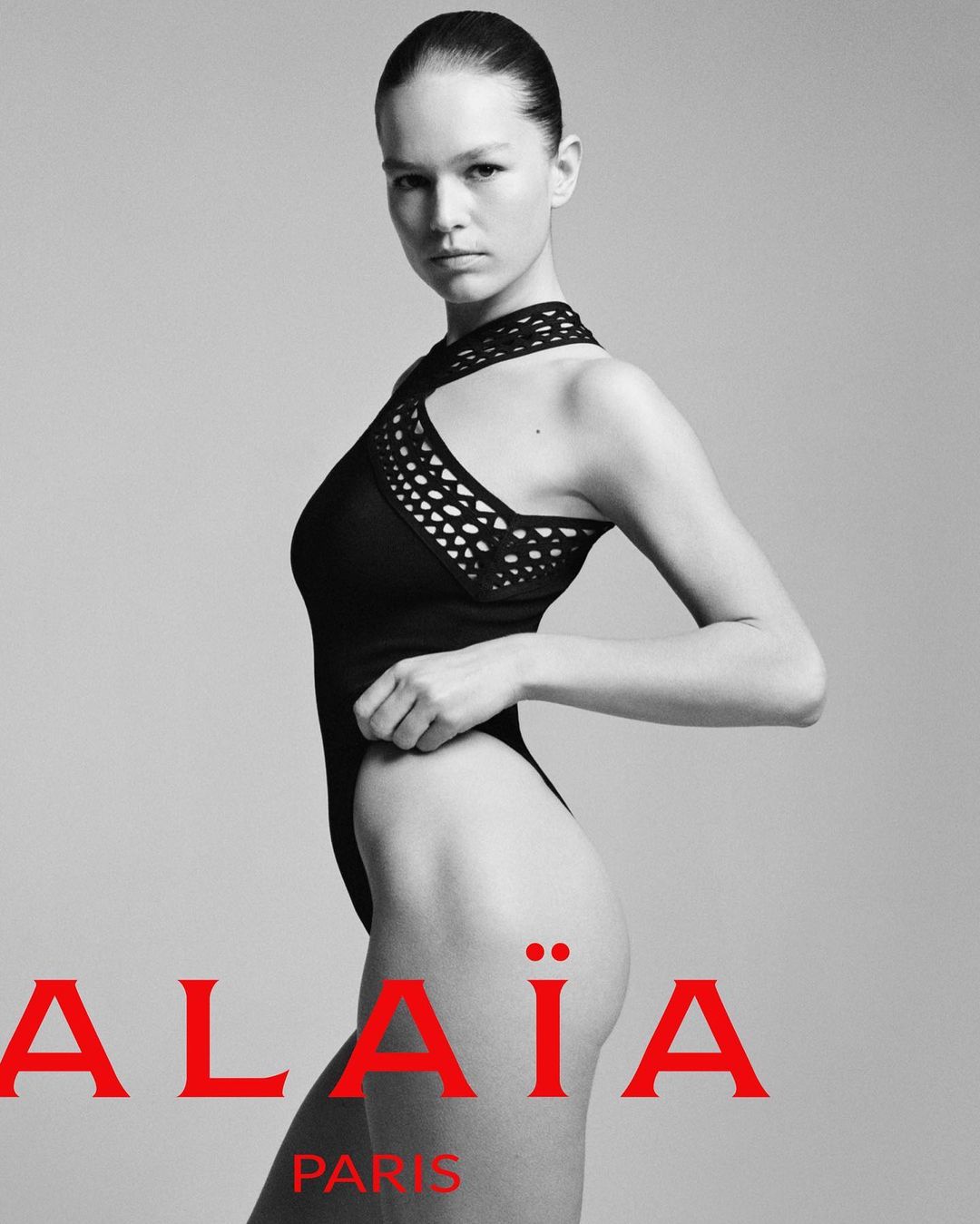 Fashion shot of a model in a black one-piece swimsuit, posing in front of a grey studio backdrop, featuring the brand name ALAÏA.