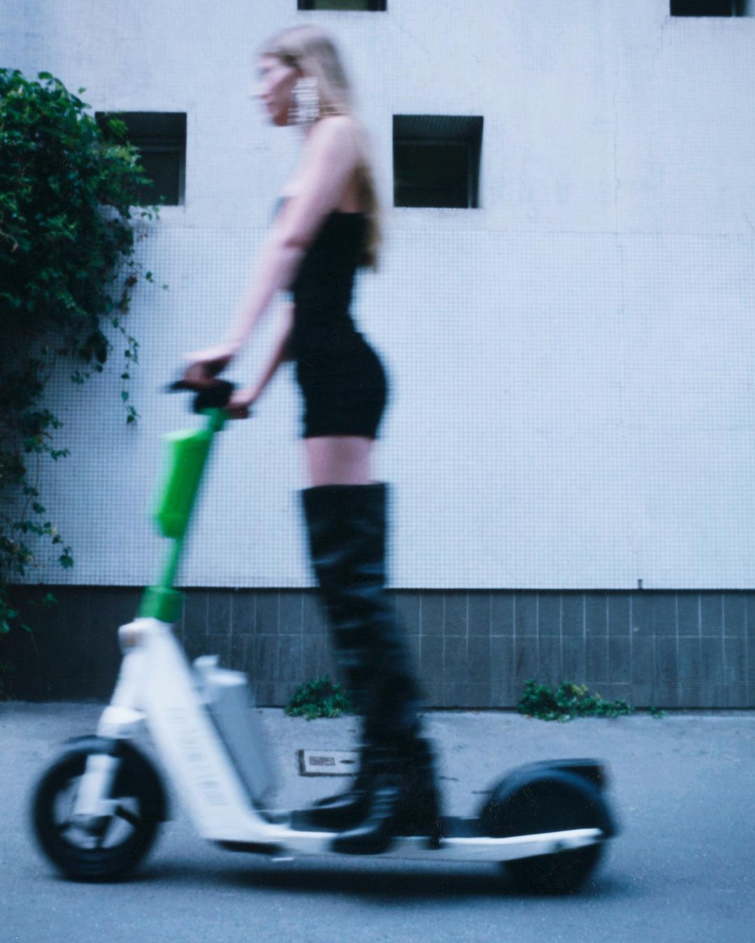 Blurred shot of a woman in a black dress and boots riding an electric scooter on a city street near a white building.