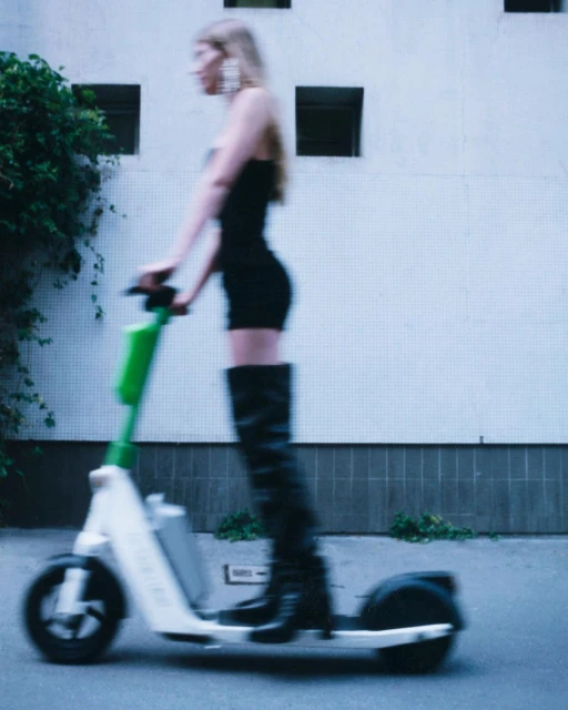 Blurred shot of a woman in a black dress and boots riding an electric scooter on a city street near a white building.