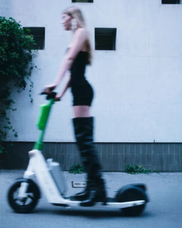 Blurred shot of a woman in a black dress and boots riding an electric scooter on a city street near a white building.