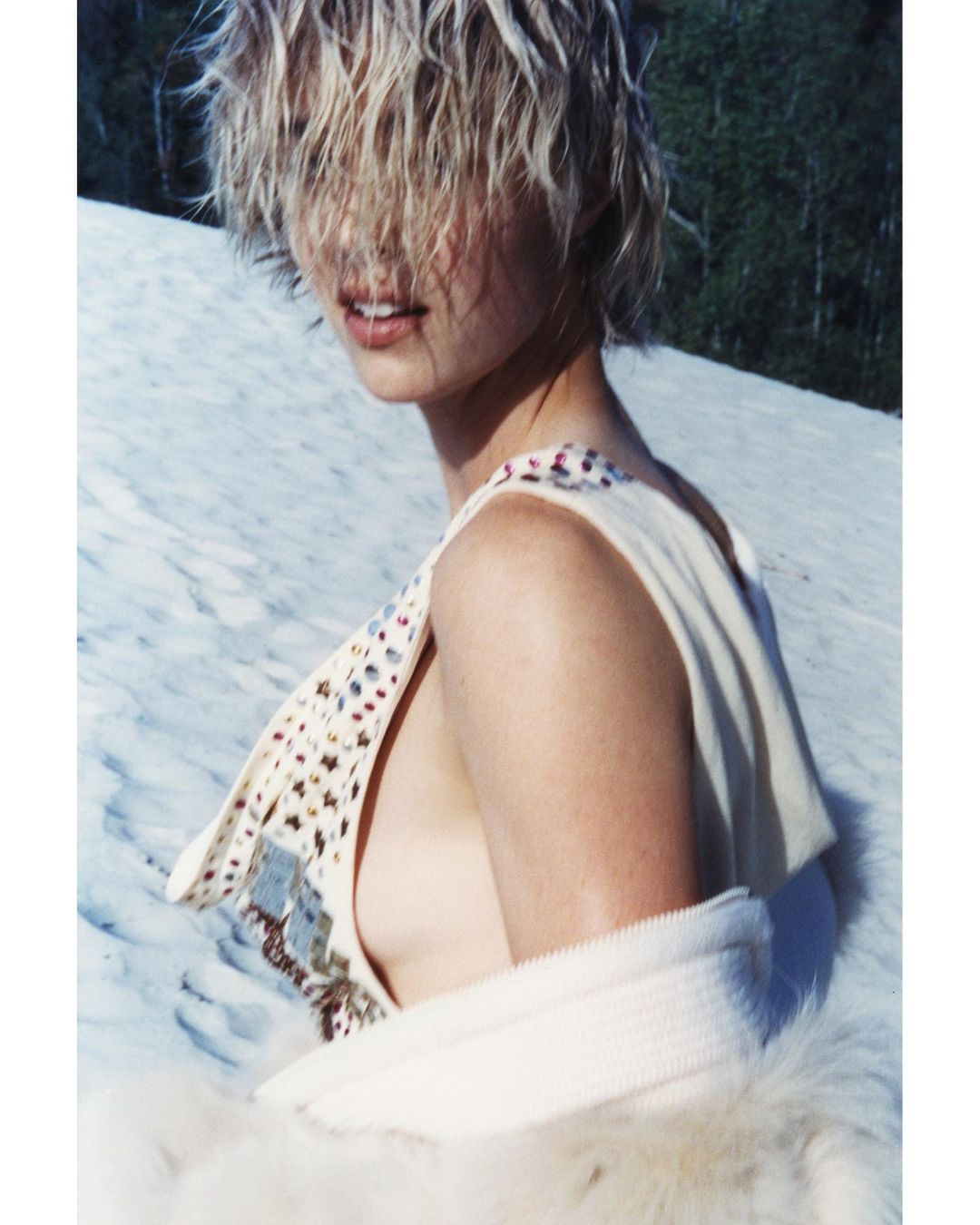 A woman poses on snow, wearing a cream-colored dress and a fluffy white stole. Her wet, blond hair falls over her face in sunlight.
