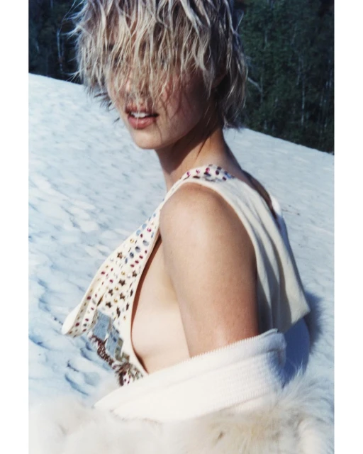 A woman poses on snow, wearing a cream-colored dress and a fluffy white stole. Her wet, blond hair falls over her face in sunlight.