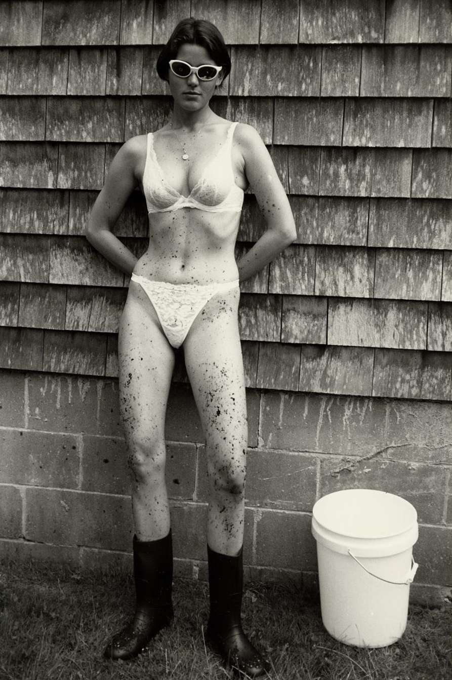 An adult woman in a bra, underwear, and boots poses against a wood-shingle wall with a bucket outside on green grass in a vintage photograph.