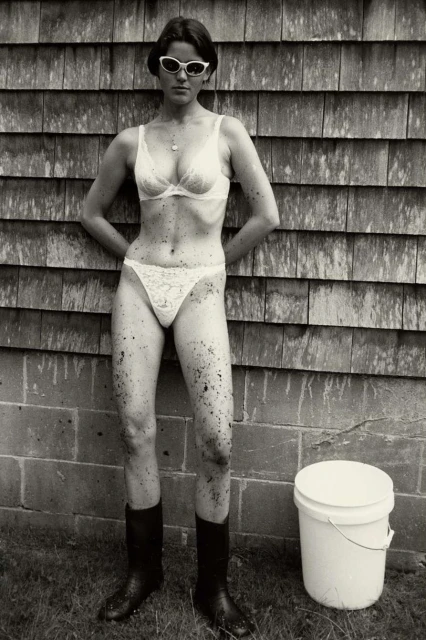 An adult woman in a bra, underwear, and boots poses against a wood-shingle wall with a bucket outside on green grass in a vintage photograph.