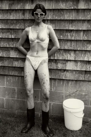An adult woman in a bra, underwear, and boots poses against a wood-shingle wall with a bucket outside on green grass in a vintage photograph.