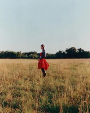 Model wearing a vibrant red skirt standing in a wide grassy field under an overcast sky.