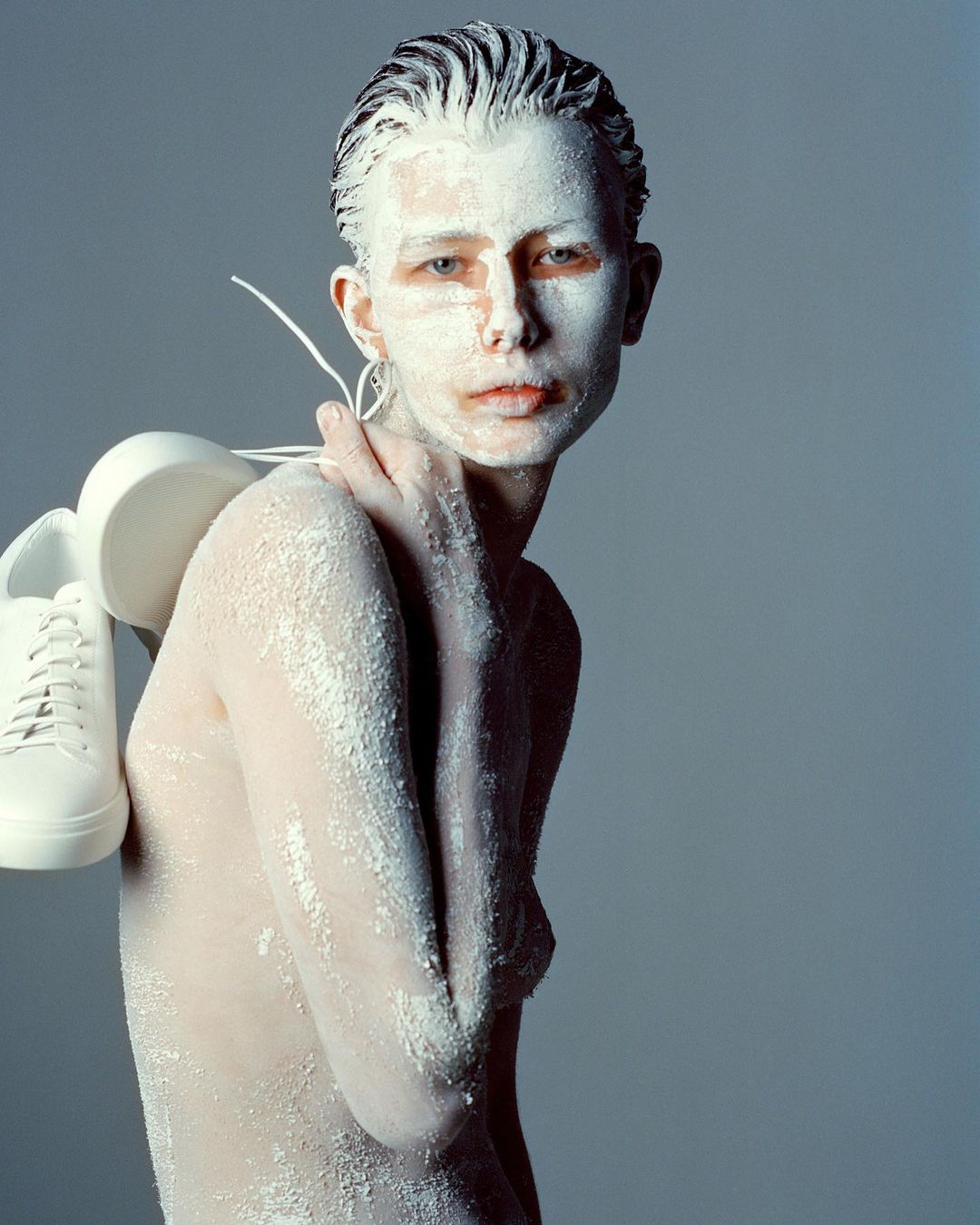 Person with white paint on their face and body holds white sneakers on their shoulder against a grey background in a studio shot.