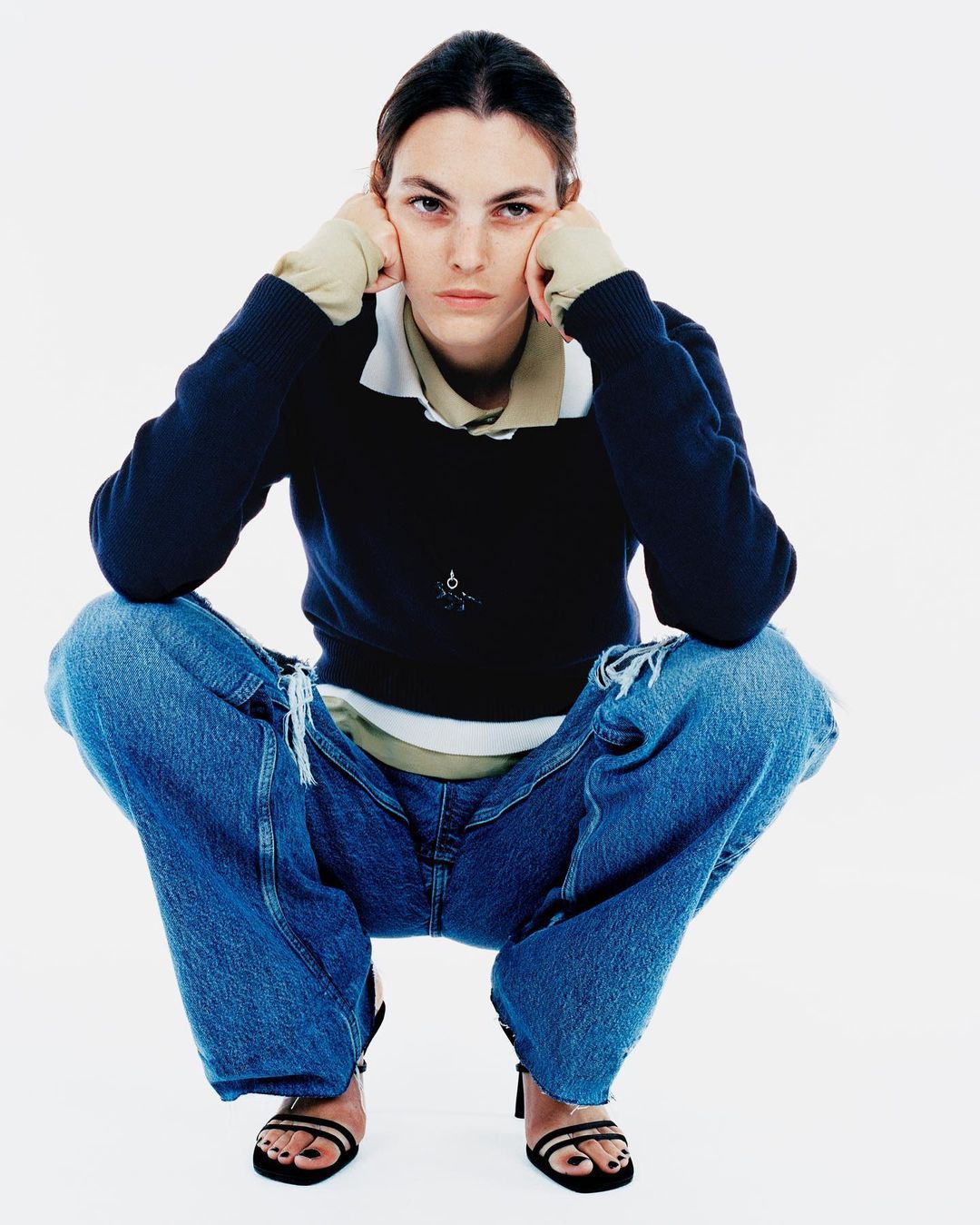 A woman in a dark blue sweater and ripped blue jeans sits in a full shot against a white backdrop, looking directly at the camera.