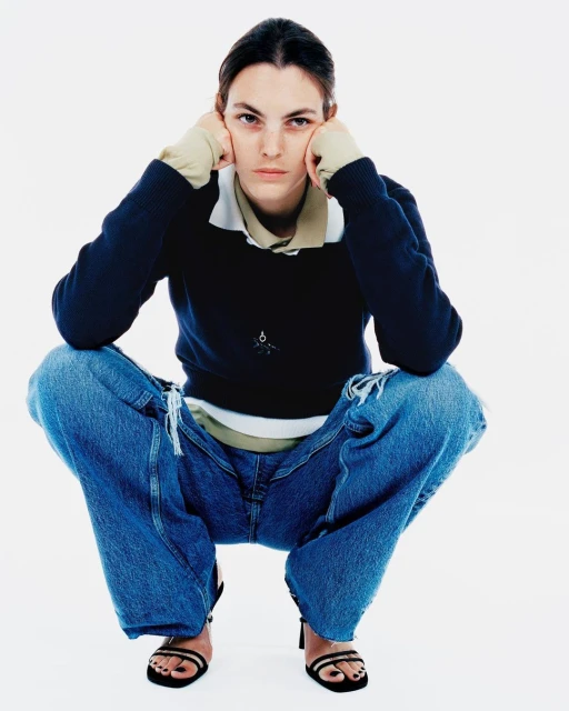 A woman in a dark blue sweater and ripped blue jeans sits in a full shot against a white backdrop, looking directly at the camera.