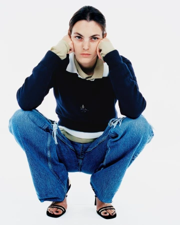 A woman in a dark blue sweater and ripped blue jeans sits in a full shot against a white backdrop, looking directly at the camera.