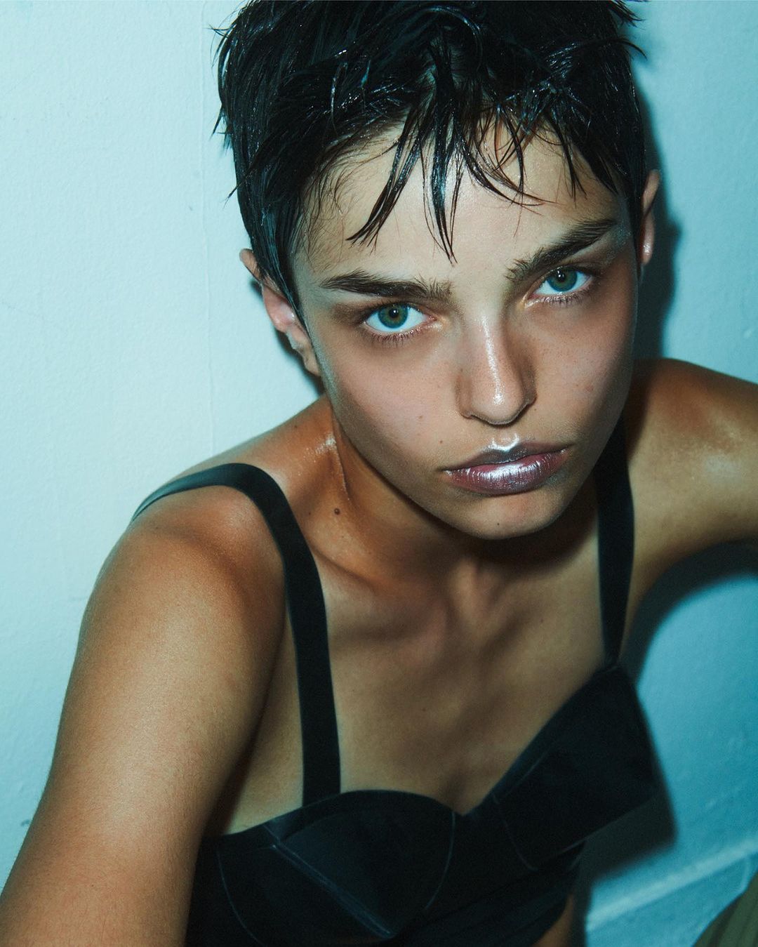 Close-up studio shot of a young adult with slicked-back dark hair, wearing a black top, highlighted lips, and standing against a light blue background.