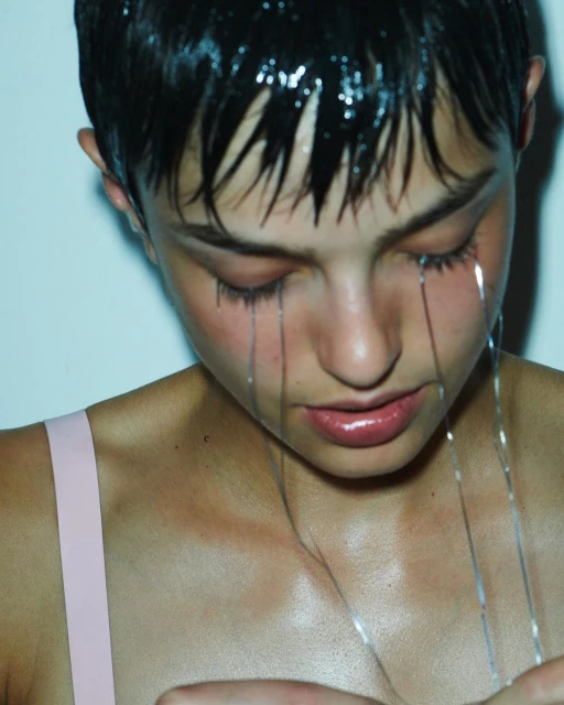 Close-up of a woman with wet dark hair, and shiny liquid running from her hairline down her face and neck, against a white background.