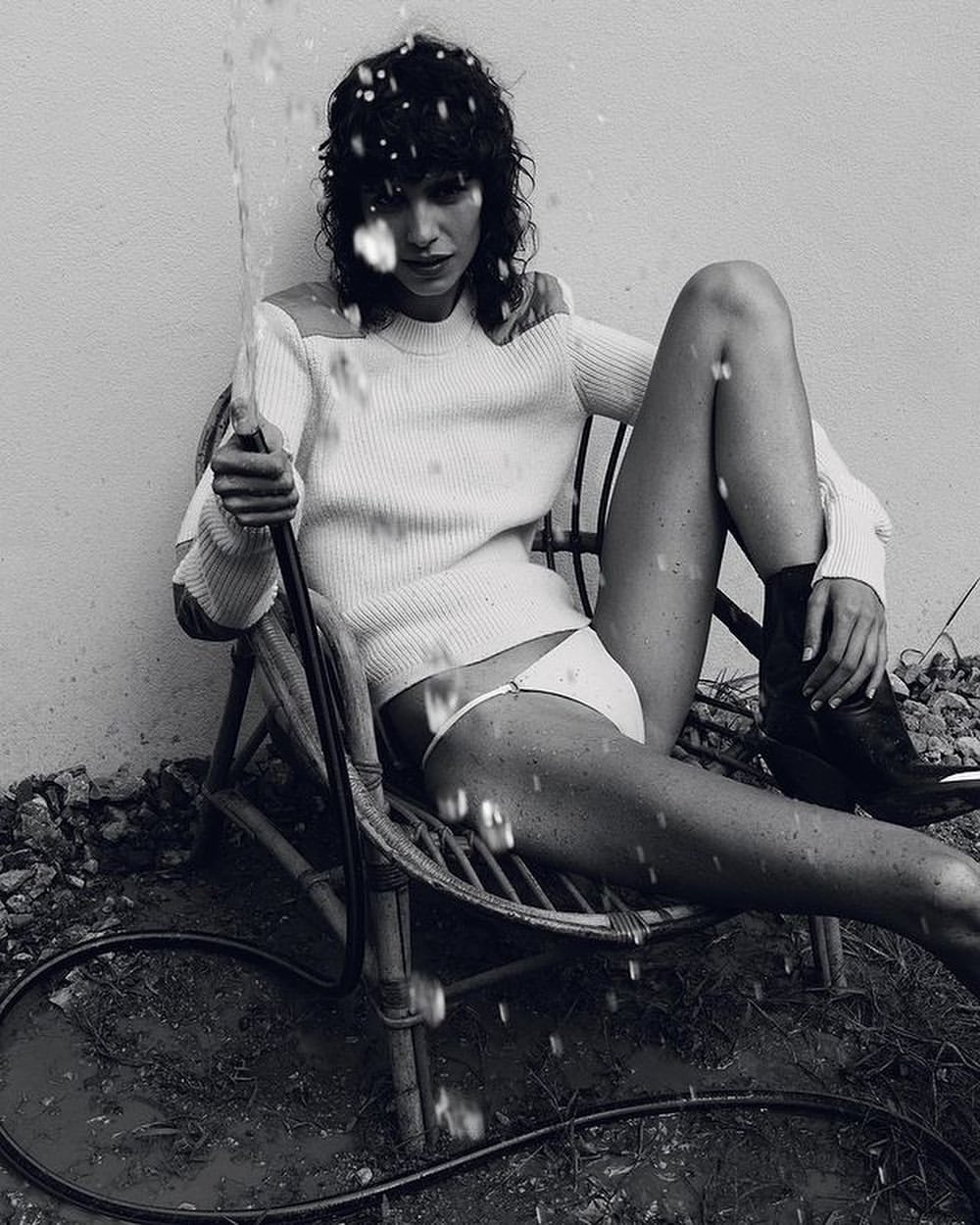 Monochrome portrait of a woman in a chair holding a hose, water spraying, wearing a white sweater and bikini bottoms, dark boots.