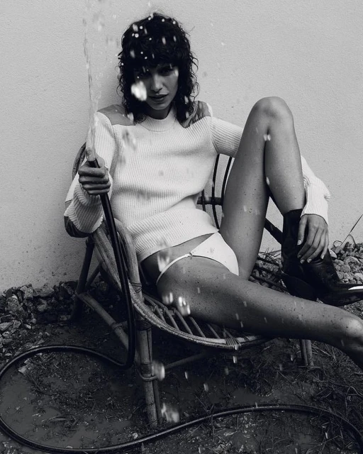 Monochrome portrait of a woman in a chair holding a hose, water spraying, wearing a white sweater and bikini bottoms, dark boots.