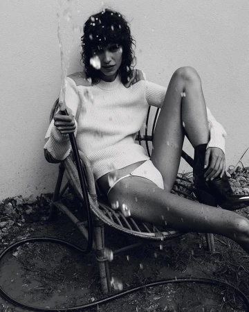 Monochrome portrait of a woman in a chair holding a hose, water spraying, wearing a white sweater and bikini bottoms, dark boots.