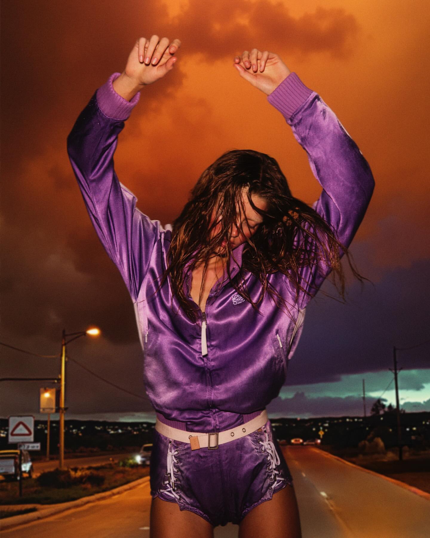 Woman in purple jacket dances on road at dusk. Orange clouds fill the sky. Streetlights and distant city lights glow.