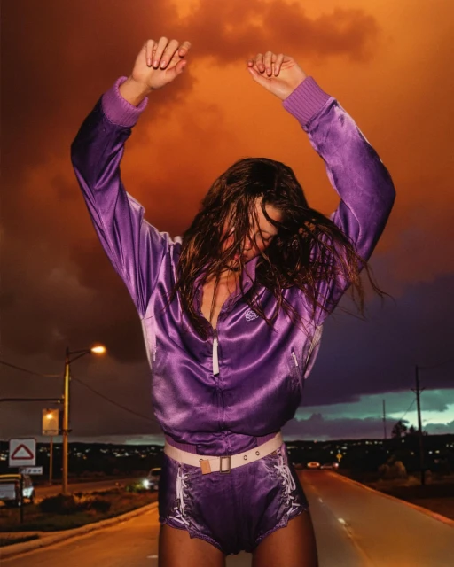 Woman in purple jacket dances on road at dusk. Orange clouds fill the sky. Streetlights and distant city lights glow.