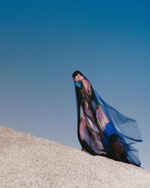 Woman in a flowing veil ascends a bright, sandy slope against a clear blue sky, creating a serene and minimalist fashion portrait.