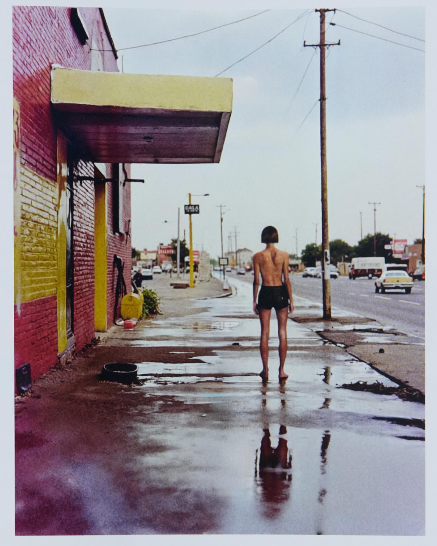 Barefoot woman stands on a wet sidewalk near a brick building and busy road with passing vehicles reflecting in the puddles.