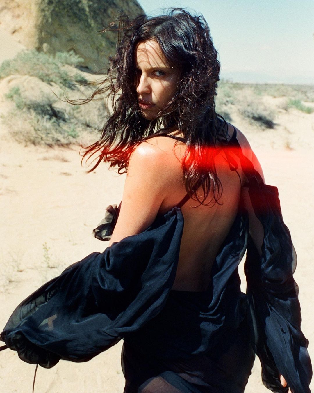 Woman with dark hair and wearing a black sheer garment posing in a desert landscape looking back over her shoulder with a serious facial expression.