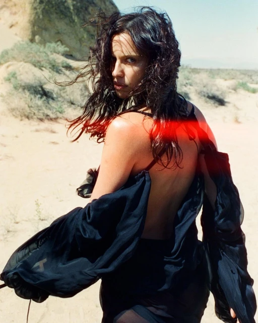 Woman with dark hair and wearing a black sheer garment posing in a desert landscape looking back over her shoulder with a serious facial expression.