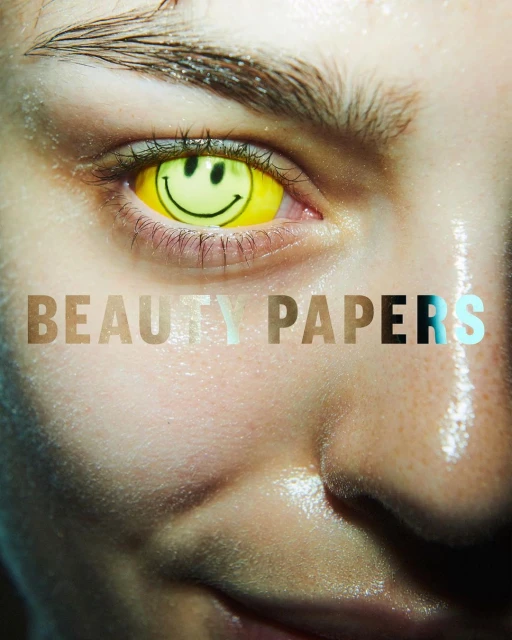 Close-up of a person's face featuring a yellow smiley face contact lens. The image also features the words BEAUTY PAPERS.