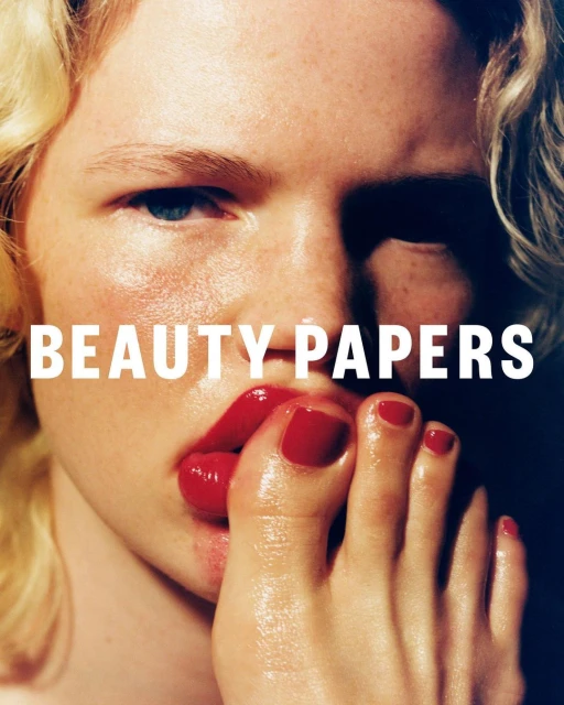 Close-up of a fair-skinned person with blonde hair and red lips, putting their foot in their mouth. The image also includes text: BEAUTY PAPERS.