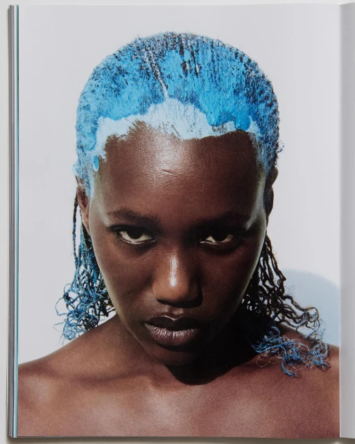 Close-up portrait of a woman with dark skin and blue dyed hair in a studio setting. The image is brightly lit, highlighting her features.