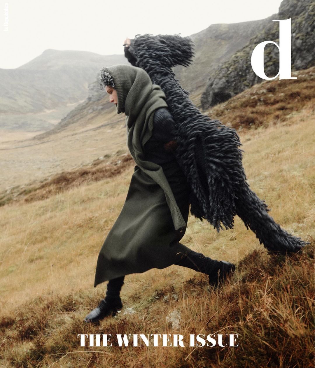 Fashionable adult strides down grassy slope carrying oversized black coat against backdrop of misty mountains; magazine cover look.