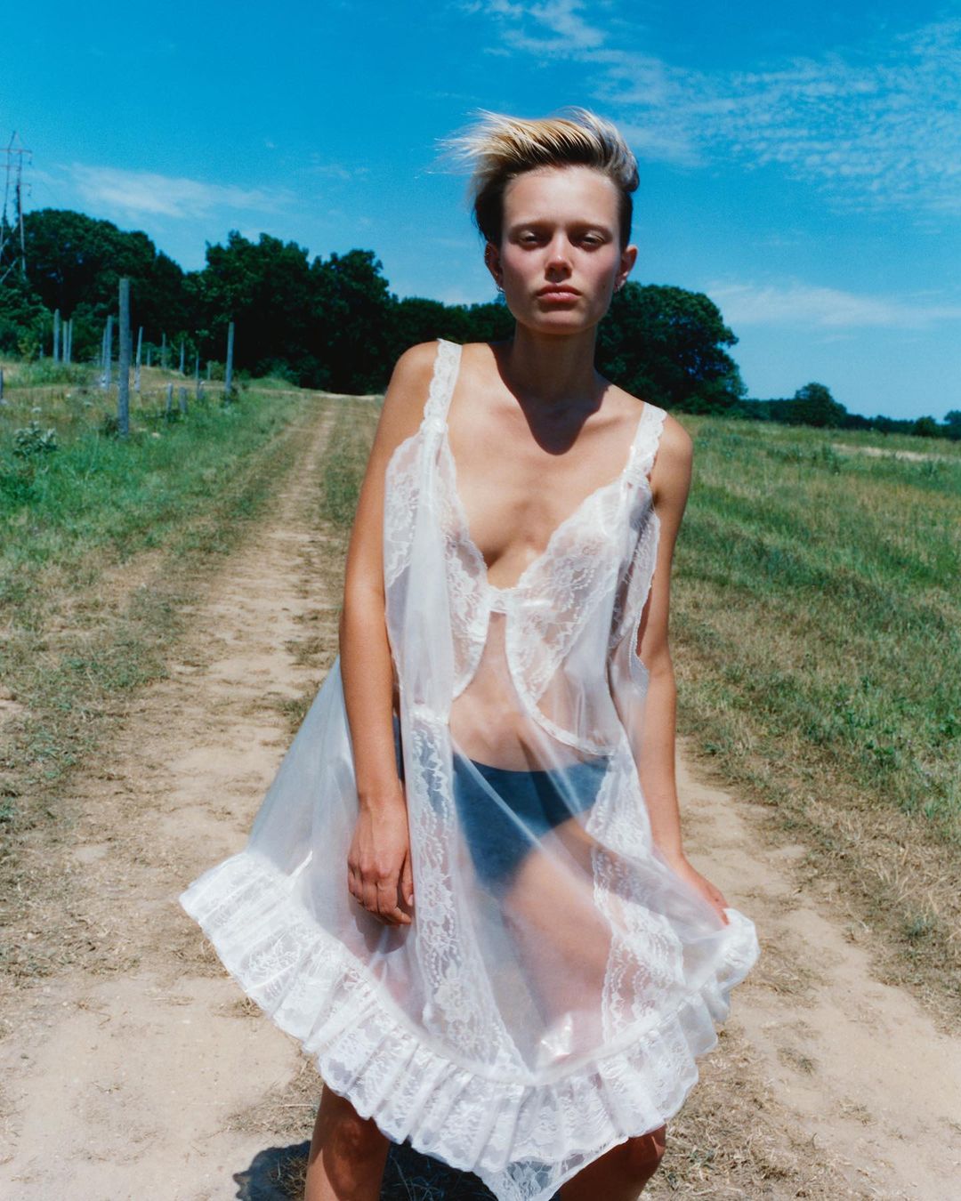 Woman in a semi-sheer white lace nightgown poses on a grassy path under a sunny blue sky, with fields and trees in the background.