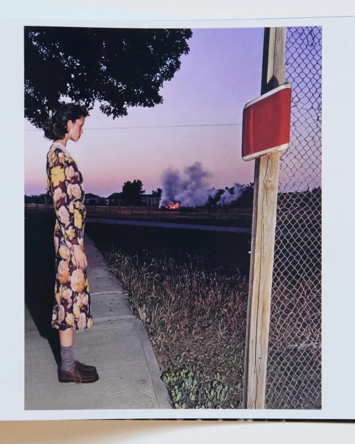 Woman in floral dress stands near a fence at dusk, watching smoke rise in the distance; roadside scene with grass and trees.